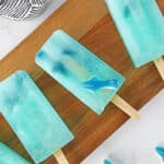 Three blue shark popsicles with gummy shark candies inside are arranged on a wooden board, with part of a black-and-white cloth visible in the corner.