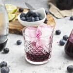 A glass with large purple-tinted ice cubes and juice sits on a table with blueberries, a small pitcher of juice, sparkling water, lime wedges, and a bowl of blueberries—perfect for festive Christmas brunch ideas.
