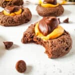 A close-up of buckeye cookies topped with creamy peanut butter and a dollop of chocolate. One cookie has a bite taken out, and chocolate chips are scattered on the white surface around the cookies.