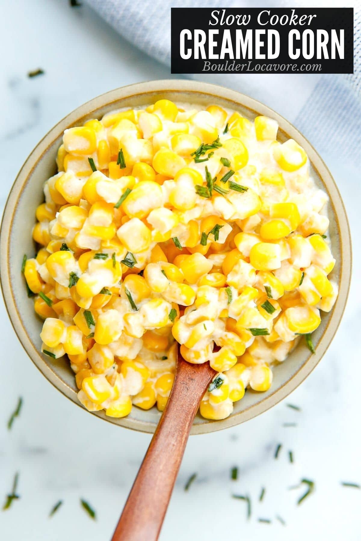 A bowl of creamy yellow and white corn topped with chopped chives, with a wooden spoon inside. Text at the top reads Slow Cooker Creamed Corn, BoulderLocavore.com.