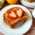 Two slices of French toast stacked on a white plate, topped with two strawberry halves, showcase classic french toast recipes. A small pitcher of syrup, orange juice, and a striped napkin sit nearby on the wooden table.