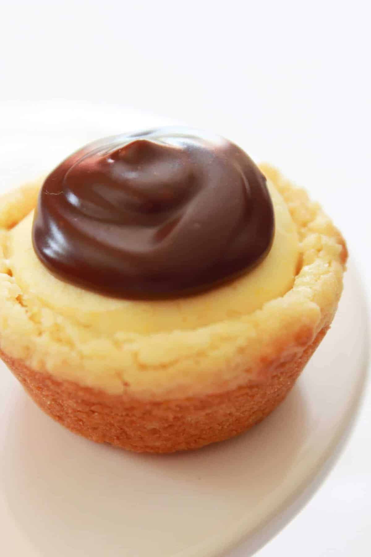 A close-up of a Boston cream pie cupcake topped with creamy yellow custard and a swirl of glossy chocolate ganache, sitting on a white plate—perfect inspiration for mini dessert recipes.