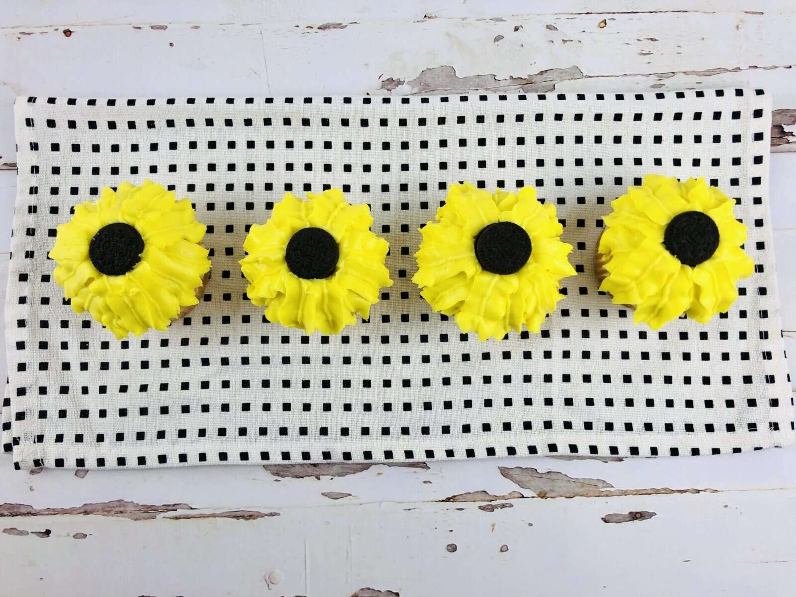 Four sunflower cupcakes with bright yellow petals and dark centers are arranged in a row on a black-and-white checkered cloth atop a rustic white wooden surface.
