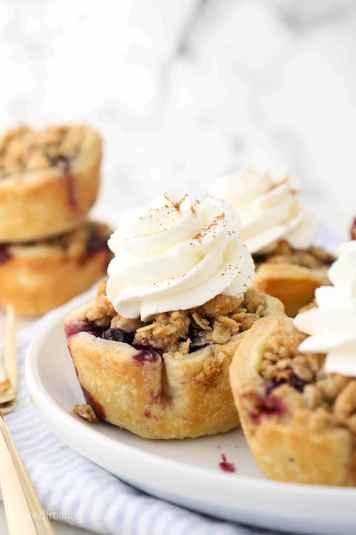 Mini fruit pies, perfect as mini desserts, are topped with whipped cream and a sprinkle of cinnamon on a white plate. Each features a crumbly topping and golden crust, with hints of berry filling peeking around the edges.