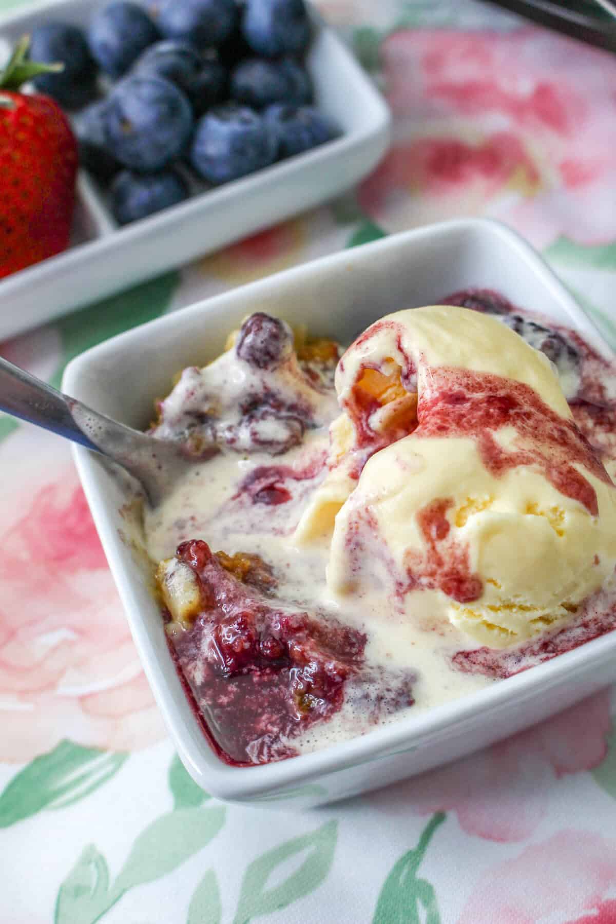 A dish of mixed berry cobbler topped with vanilla ice cream next to a slow cooker and fresh berries.