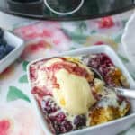 A dish of mixed berry cobbler topped with vanilla ice cream next to a slow cooker and fresh berries.