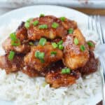 A plate of white rice topped with sweet and sour chicken—glazed, crispy pieces garnished with chopped green onions, with a fork and knife placed on the side of the plate.