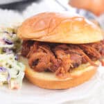 A pulled pork sandwich on a soft bun sits on a white plate next to creamy coleslaw, one of the classic sides for pulled pork sandwiches.