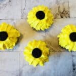 Four cupcakes, made with yellow cake mix and decorated to look like sunflowers, feature bright yellow frosting petals and a chocolate cookie center, arranged in a diamond shape on a light marbled surface.