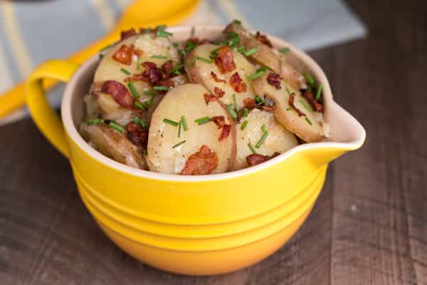 A yellow bowl filled with sliced potatoes topped with bacon pieces and chopped chives, sitting on a wooden table—perfect for adding to your list of 4th of July crock pot recipes.