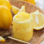 A small glass jar filled with creamy lemon curd sits in a wicker basket, surrounded by whole and sliced fresh lemons—perfect inspiration for Yellow Food Board Ideas. A silver spoon rests nearby.