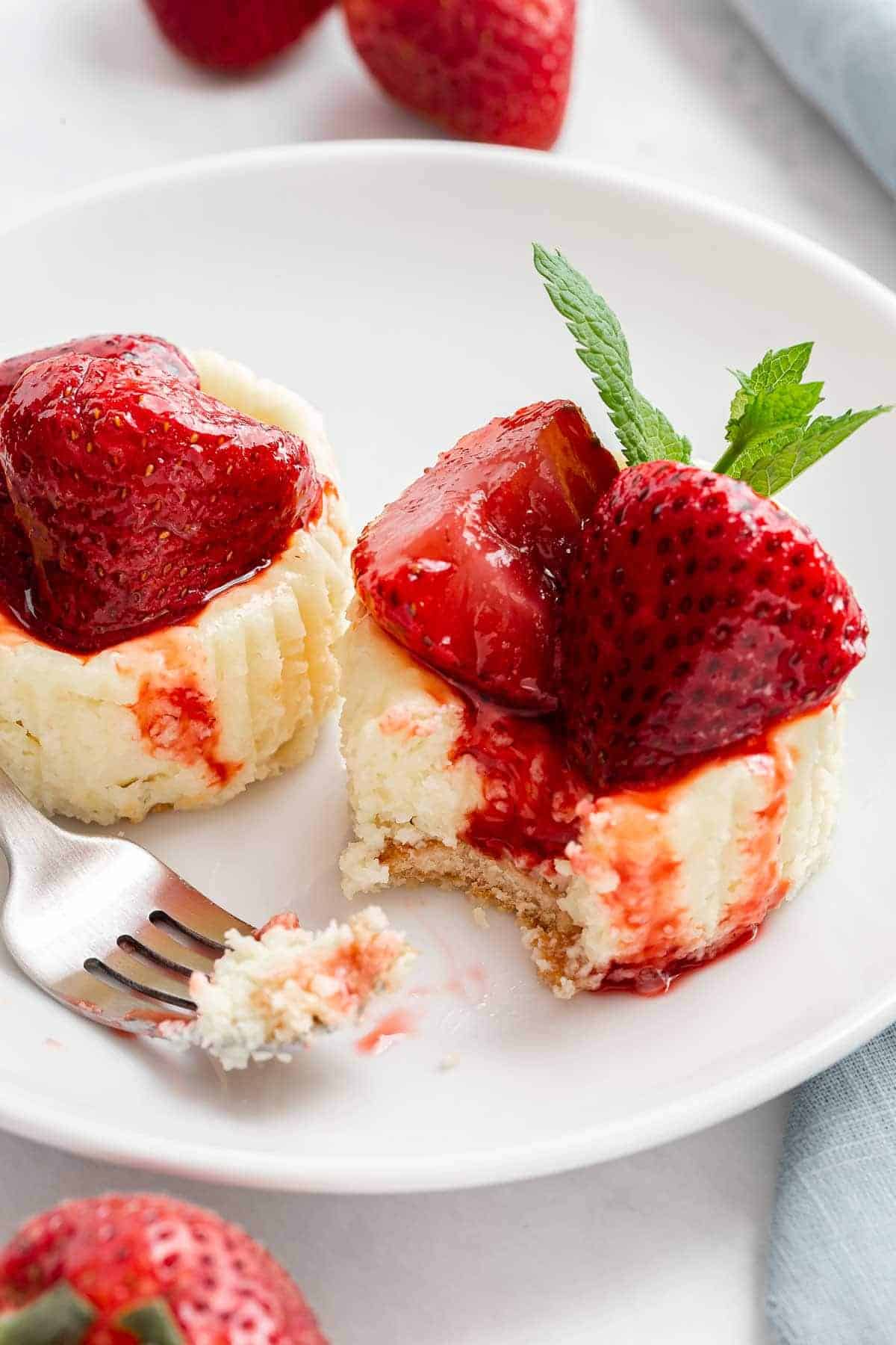 A delicious mini dessert, this small cheesecake is topped with glazed strawberries and a mint leaf on a white plate. A fork reveals its creamy texture and crumbly crust, while fresh strawberries add the perfect finishing touch.