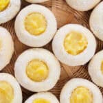 Round Lemon Curd Cookies with a yellow filling in the center are arranged closely together on a woven bamboo tray. The pastries have a light dusting of flour on their surfaces.