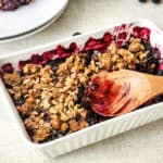 A white baking dish filled with blueberry crumble, partially served, with a wooden spoon resting on top. Some of the crumble and blueberries are visible, evoking the rustic charm of homemade berry pies.