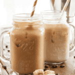 Two mason jar mugs filled with iced coffee and striped straws sit on a wooden table scattered with coffee beans, ice cubes, and brown sugar cubes. A metal pot in the background hints at how to make the best iced coffee from home.