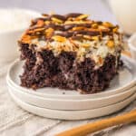 A slice of chocolate poke cake topped with toasted almonds and shredded coconut sits on stacked white plates, with a wooden spoon and small bowls in the background.