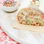 A slice of vanilla funfetti cake roll with colorful sprinkles, topped with white frosting and more sprinkles, is displayed on a wooden board next to a small bowl of rainbow sprinkles—an irresistible vanilla funfetti cake roll recipe in every bite.