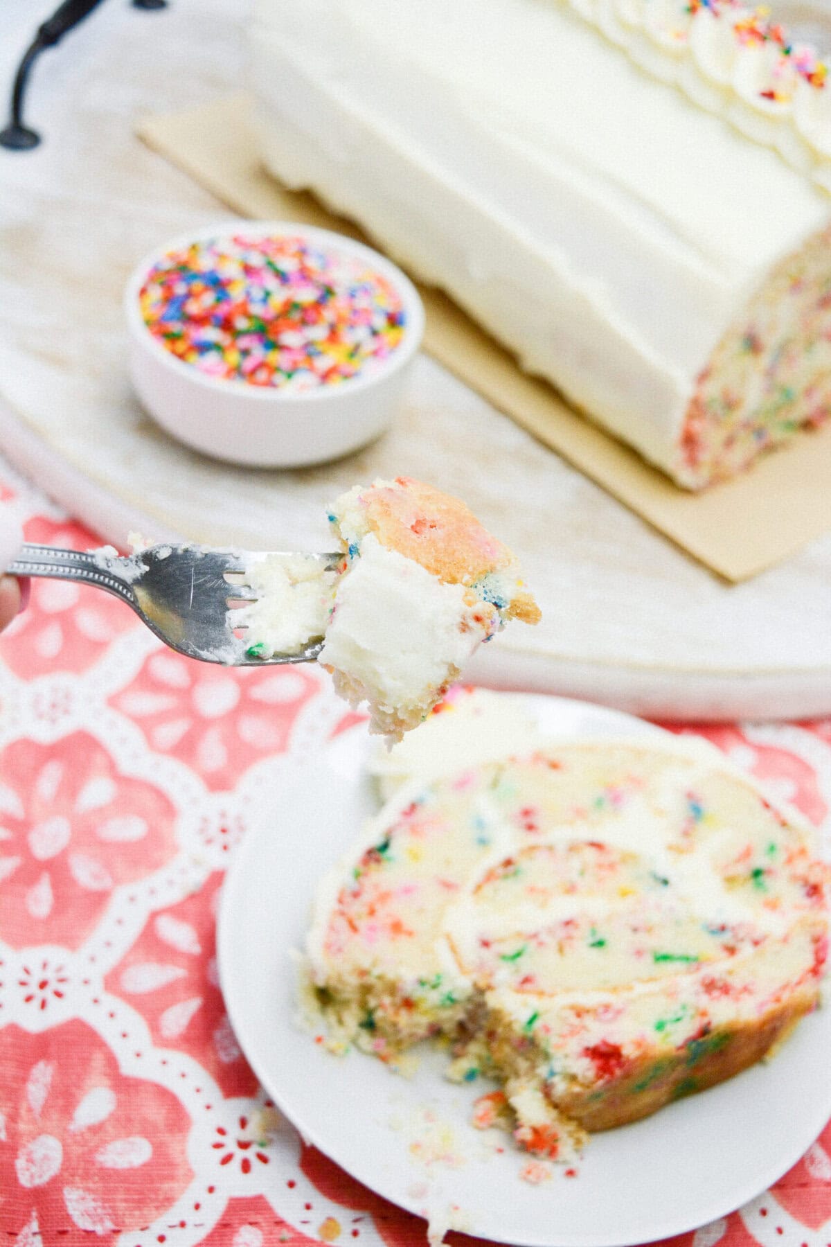 A fork holds a bite of vanilla funfetti cake with white frosting above a plate with a slice of the same cake. A bowl of colorful sprinkles and the remaining vanilla funfetti cake roll recipe are in the background on a white surface.