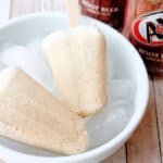 Two creamy root beer popsicles on wooden sticks rest on ice cubes in a white bowl, with an A&W root beer can in the background—perfect summer snacks for kids on a hot day.