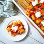 A white plate holds a cheesy serving from this recipe for tater tot casserole with sour cream, bacon, chopped tomatoes, and green onions. Nearby, a baking dish with extra casserole and a wooden spatula rests on a white wooden table.