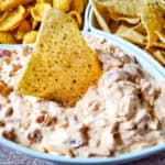 A close-up of a bowl of creamy football dip, topped with chopped ingredients and a tortilla chip. Bowls of corn chips and tortilla chips are in the background—perfect for fans searching for delicious football dip recipes.