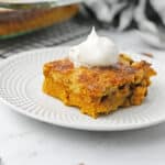 A slice of pumpkin dessert topped with whipped cream sits on a white plate. Cinnamon sticks and pecans are scattered nearby, hinting at flavors found in Easy Thanksgiving Dessert Recipes, with a baking dish and striped cloth in the background.
