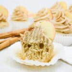 A group of frosted cupcakes topped with apple slices sits on a white surface. One cupcake in front has a bite taken out, revealing its soft interior—perfect for those seeking easy Thanksgiving dessert recipes. Two cinnamon sticks lie beside the cupcakes.