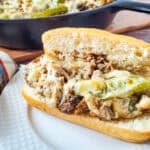A hoagie roll filled with melted cheese, sliced beef, onions, and green peppers sits on a white plate. In the background, a cast iron skillet with more of the sandwich filling is visible.