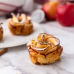 Close-up of an apple roses pastry shaped like a rose, dusted with powdered sugar. More pastries and red apples are blurred in the background on a white marble surface, with a cinnamon stick lying nearby.