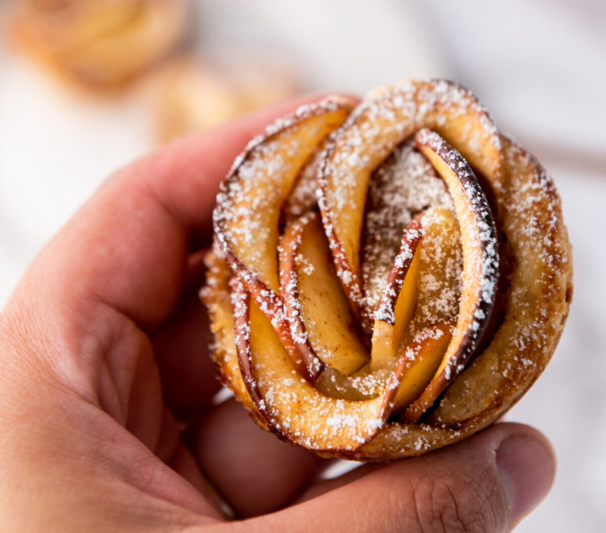 A hand holds a small pastry shaped like a rose, made with thin apple slices and dusted with powdered sugar. Blurry apples and plates are visible in the background.