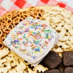 A platter with animal crackers, mini pretzels, and brownies surrounds a bowl of creamy fall dessert dip topped with colorful rainbow sprinkles, set on a red and white checkered cloth.