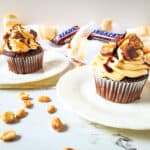 Two Snickers Cupcakes with caramel-colored frosting, topped with chocolate drizzle, peanuts, and chopped candy bars, sit on white plates. Peanuts and candy bars are scattered on a white wooden table with a yellow towel in the background.