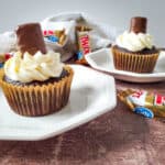 Indulge in decadent Twix cupcakes—two chocolate cupcakes with white frosting and a mini Twix bar on top, served on white plates with Twix candy wrappers in the background on a wooden surface.