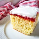 A slice of yellow sponge cake from our favorite 9x13 cake recipes is topped with red fruit jam and fluffy whipped cream, served on a white plate with a red and white patterned cloth in the background.