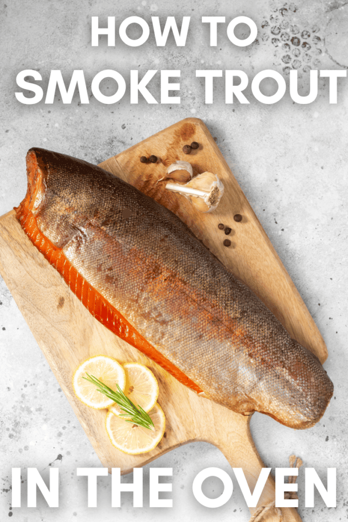 A whole smoked trout on a wooden cutting board with lemon slices, garlic, and peppercorns, on a gray surface. Large text reads: How to Smoke Trout in the Oven—a simple smoked trout recipe.
