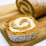 A pumpkin roll cake, reminiscent of gingerbread desserts, is dusted with powdered sugar on a wooden cutting board, with one slice cut to reveal a spiral of creamy filling.