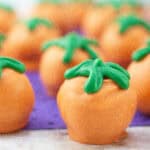 Close-up of orange candy treats shaped like pumpkins, with green frosting tops, arranged on a purple surface.
