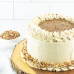 A round pecan pie cake recipe creation with white frosting, piped rosettes, a rich pecan and caramel topping in the center, and chopped pecans around the base, displayed on a wooden cake stand.