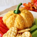A pumpkin-shaped cheese ball sits on a white plate, surrounded by sliced red bell peppers, celery sticks, and round crackers, with autumn leaves in the background.