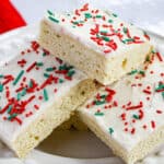 Three rectangular sugar cookie bars with white frosting and red and green sprinkles are stacked on a white plate, creating a festive holiday treat that’s perfect for enjoying with family and friends.