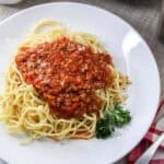 A plate of spaghetti topped with tomato meat sauce and grated cheese, garnished with a sprig of parsley, captures the spirit of an Italian Sunday Dinner. A red and white checkered napkin, fork, and black pepper shaker are beside the plate.