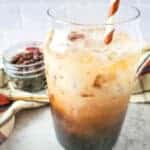 A glass of vanilla iced coffee, inspired by a Starbucks recipe, with milk swirling through it and a striped straw. In the background, a jar of coffee beans and a plaid cloth sit on a light-colored countertop.