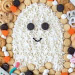 A Halloween Dessert Table features a large ghost shape made of white icing with mini cookies for eyes and mouth, surrounded by assorted cookies, marshmallows, pretzels, and festive Halloween candies on a purple tray.