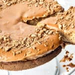 A round cheesecake with a caramel-colored topping and crushed cookie crumbs, displayed on a glass plate. Inspired by gingerbread desserts, a slice has been cut but not removed, revealing the creamy texture inside. Crumbs are scattered nearby.