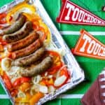 Sausage peppers and onions in oven, baked on a foil-lined tray, sit atop a football-themed tablecloth with TOUCHDOWN pennants and a football napkin nearby.