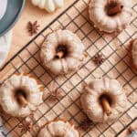 Mini bundt cakes with white icing and cinnamon sticks on top are arranged on a cooling rack, perfect for Easy Thanksgiving Dessert Recipes. Surrounded by star anise, small white pumpkins, milk, and a napkin on a wooden surface.