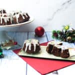 Gingerbread mini bundt cakes with white icing and colorful sprinkles are displayed on a cake stand and a plate. Holiday decorations, ornaments, and a red napkin create a festive atmosphere.