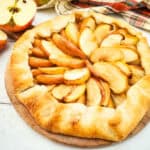 A rustic apple galette, reminiscent of a classic apple tart, with golden, flaky crust and arranged apple slices sits on a wooden board. A halved apple and a checkered cloth are nearby on a white background.