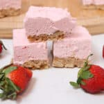 Two creamy pink strawberry dessert bars are stacked atop a crumbly crust, surrounded by fresh whole strawberries on a white surface—perfect no bake 4th of July desserts, with more bars on a wooden board in the background.
