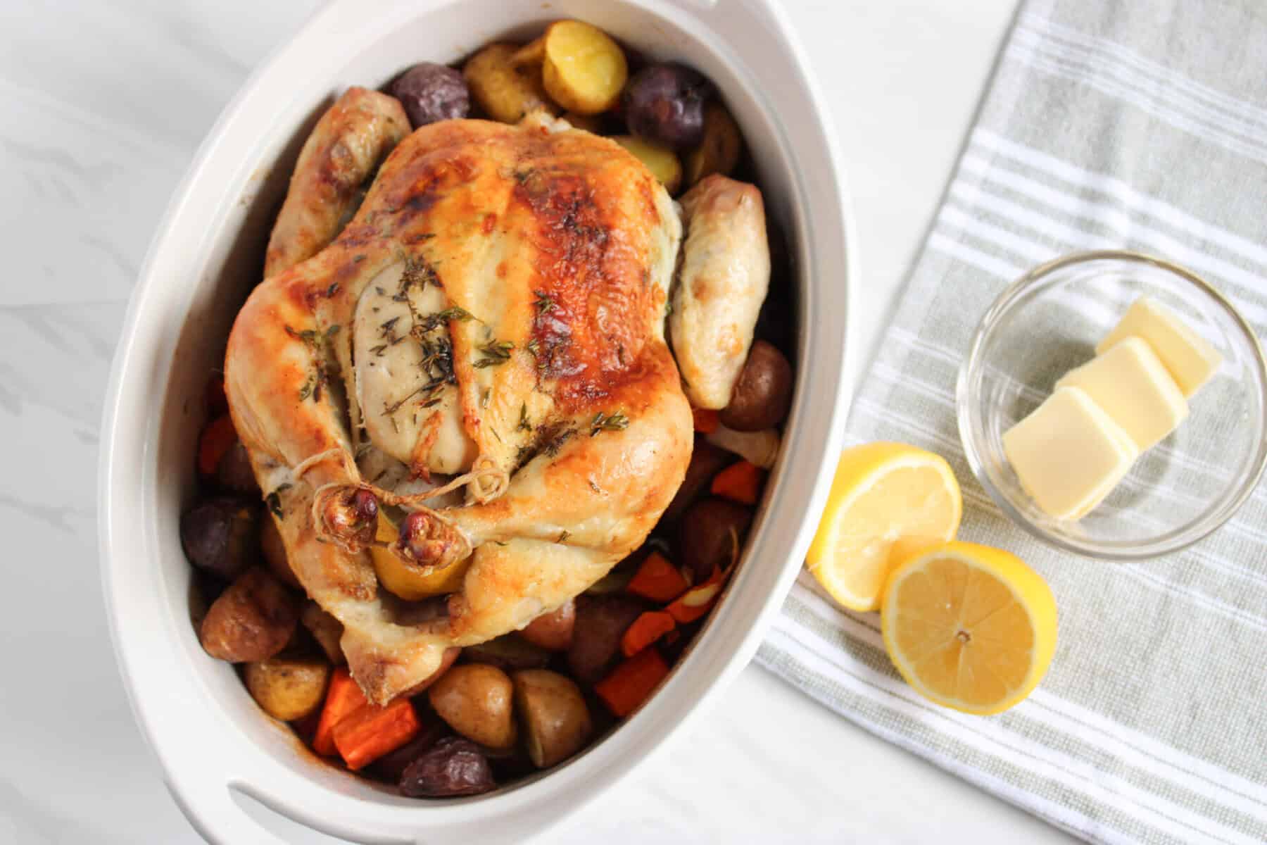 A whole baked chicken in the oven with herbs rests in a white baking dish, surrounded by potatoes and carrots. Nearby, a glass bowl holds butter slices and lemon halves beside a striped towel on a white surface.